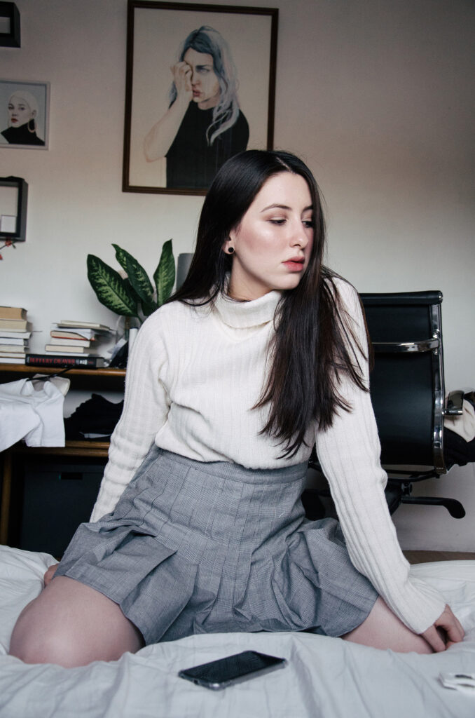 Gabriela Inzunza sits on a bed wearing a white turtleneck and gray skirt with a phone and earbuds nearby. Behind her a room is adorned with framed artworks and a potted plant.